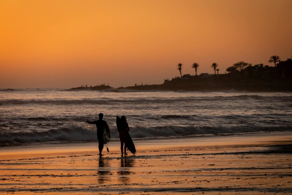 Two surfers walking along the beach at sunset in Taghazout Bay, Morocco, highlighting the coastal charm of the Africa Triathlon Cup 2025 destination.