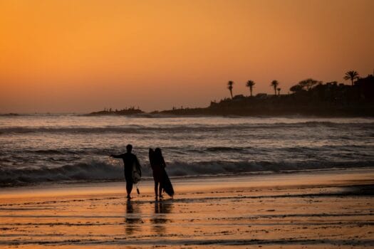 Two surfers walking along the beach at sunset in Taghazout Bay, Morocco, highlighting the coastal charm of the Africa Triathlon Cup 2025 destination.