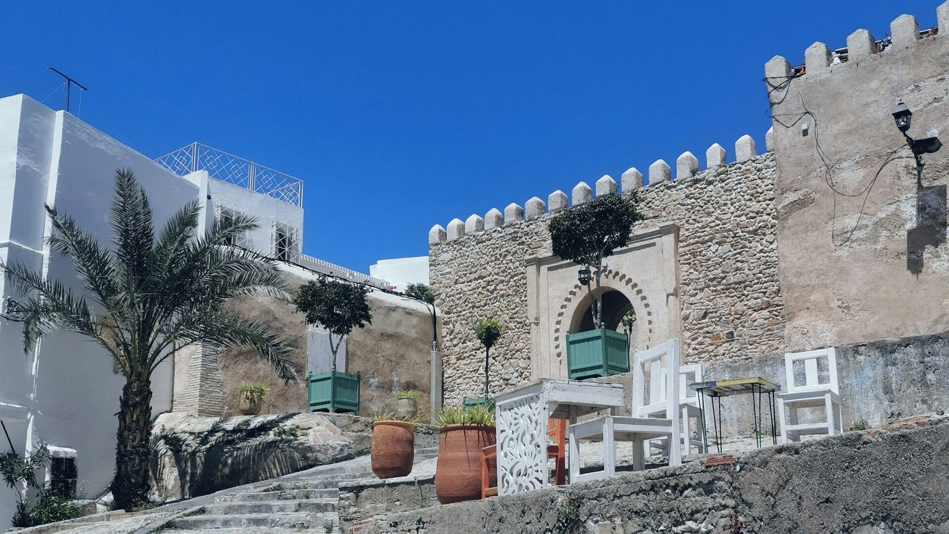 Stone alley and arched gate with white chairs and potted plants under a bright blue sky in Tangier’s Kasbah