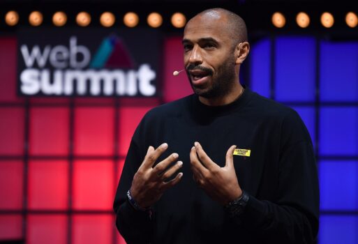 Thierry Henry speaking on stage during an event, wearing a dark suit and smiling under spotlights