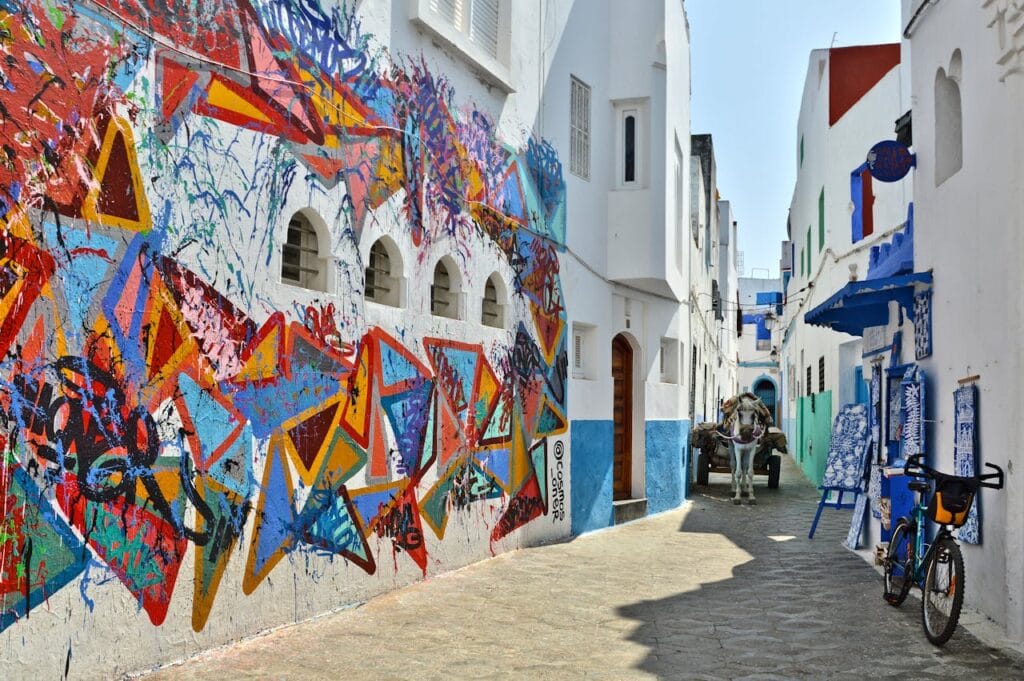 Colorful mural on white walls in Asilah Medina with a cart and bicycle nearby