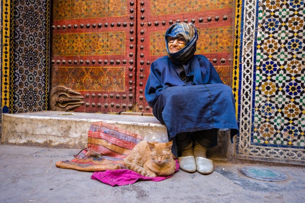 Elderly Moroccan woman sitting by a decorated door in Meknès with a cat resting beside her