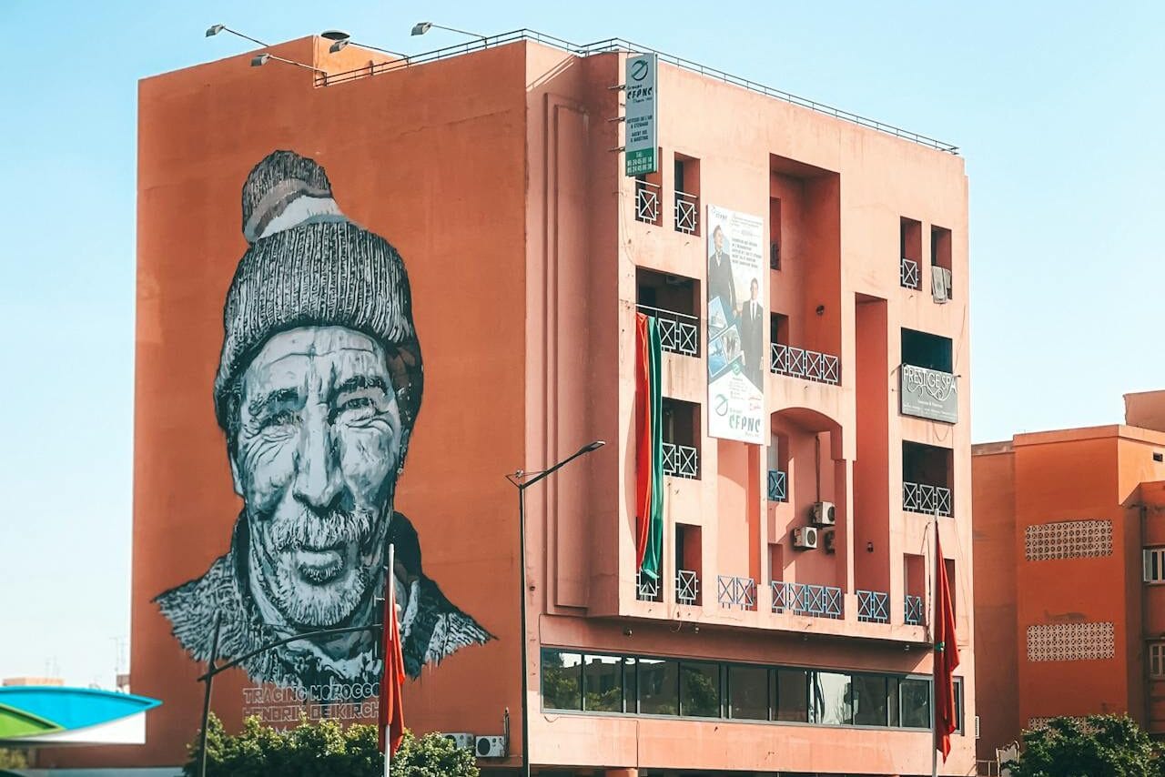 Black-and-white portrait mural painted on an orange building above a busy Moroccan street with cars and scooters