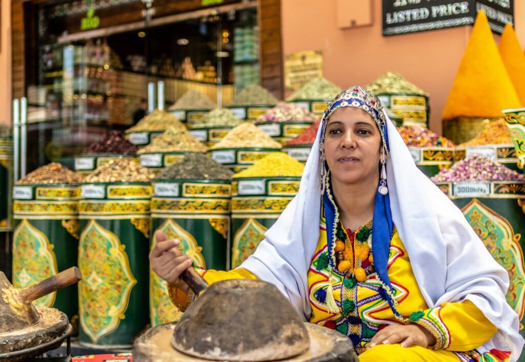 Moroccan woman in traditional Amazigh clothing preparing spices in a souk with colorful spice cones behind her.