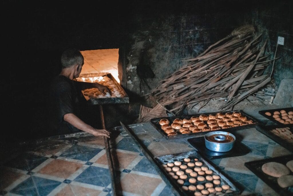 A Moroccan baker placing trays of bread and pastries into a traditional wood-fired oven inside a medina bakery.