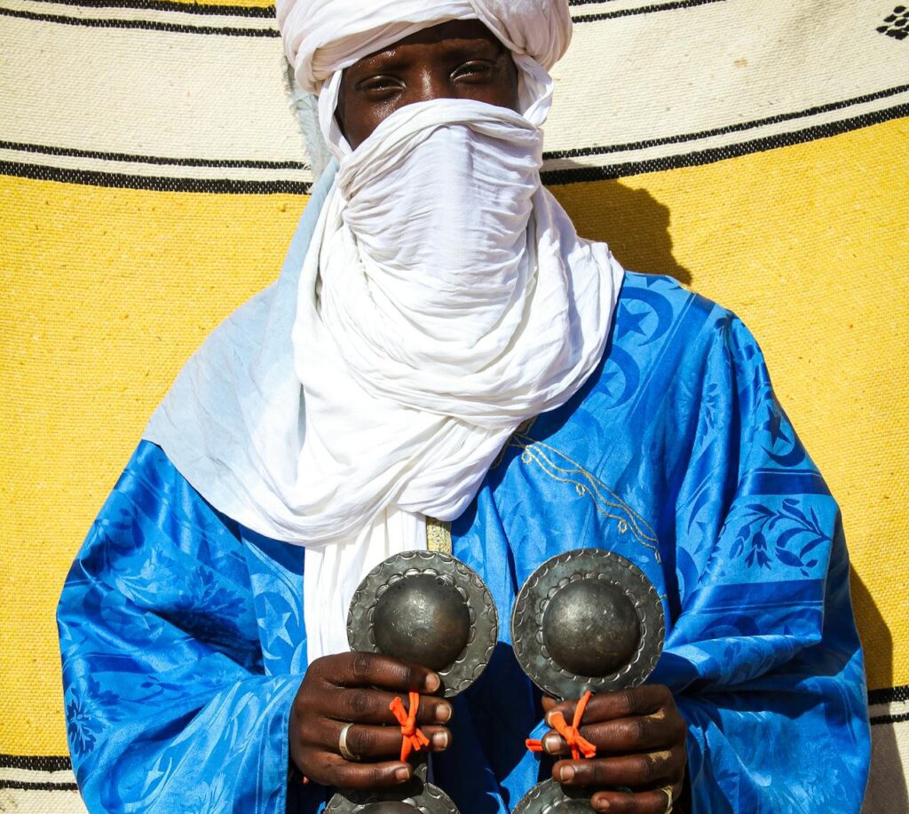 A Saharan musician wearing a blue robe and white turban holding metal castanets in the desert region of M’Hamid El Ghizlane.