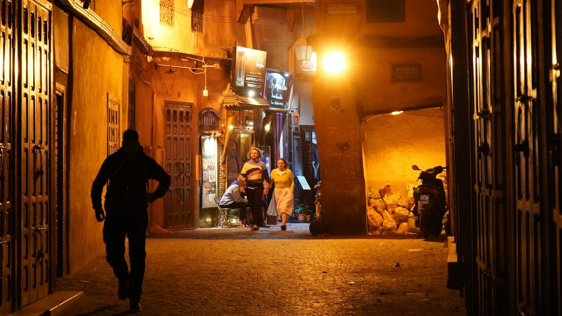 A dimly lit Marrakech medina alley at night with warm lantern light and a few people walking through the passage.