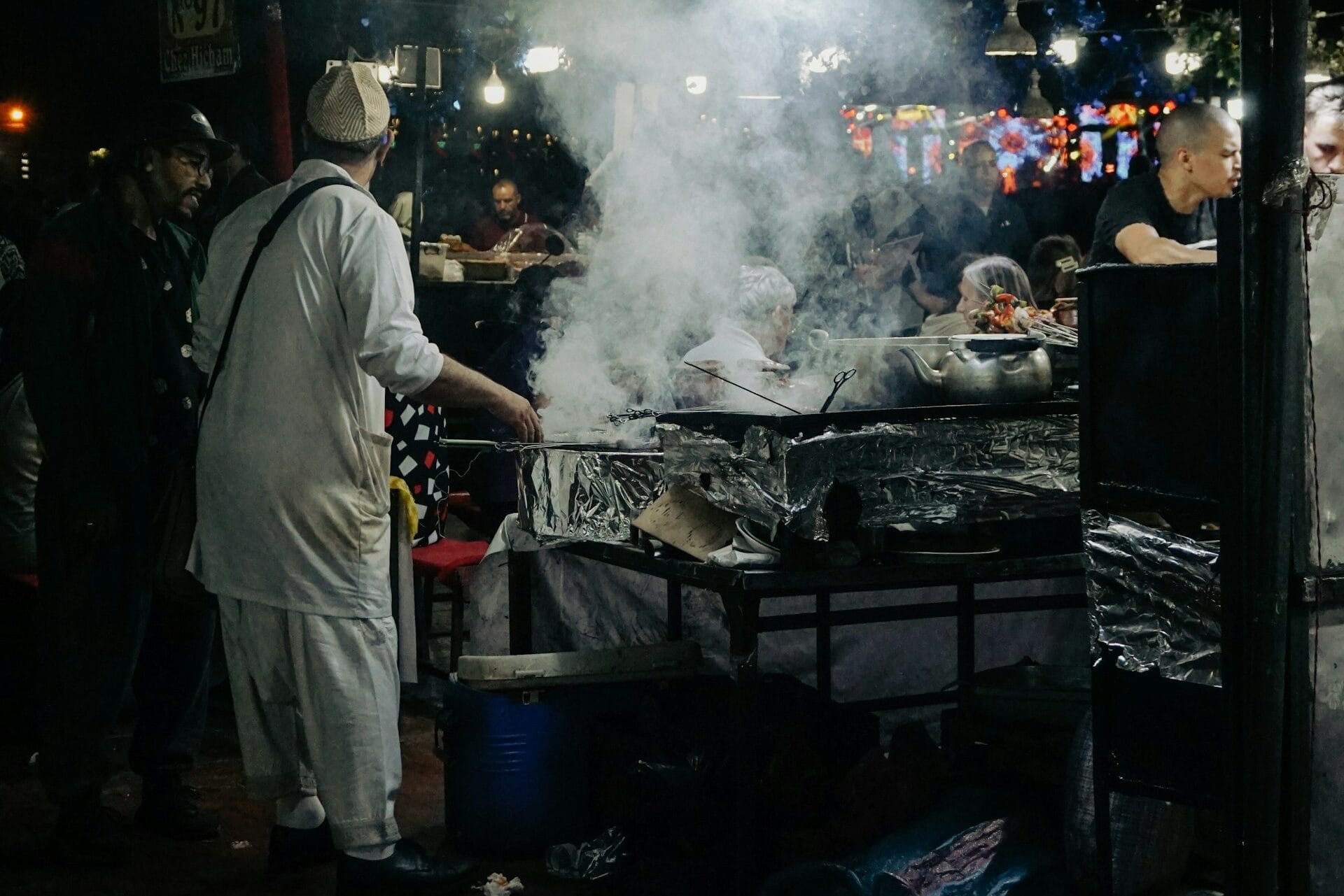 A smoky street food stall at a Marrakech night market, with vendors grilling food and people dining under dim lights