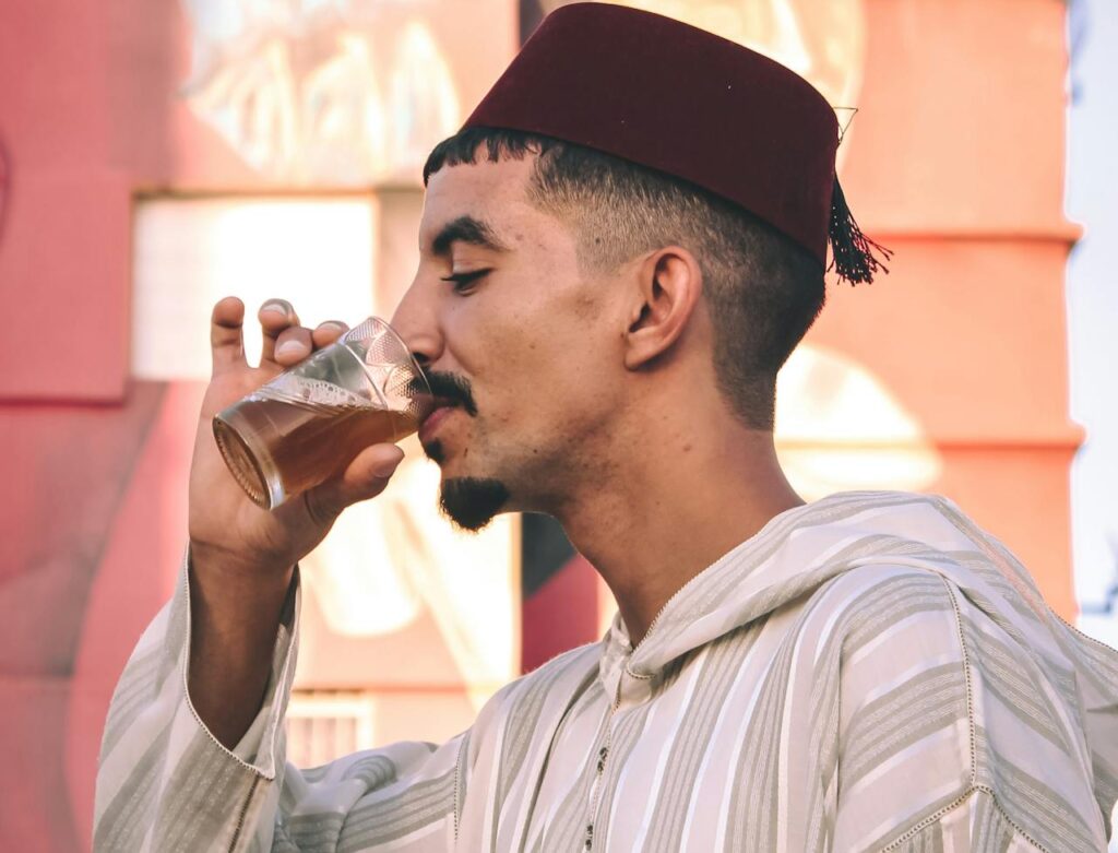 A Moroccan man in a traditional djellaba and fez hat drinking mint tea outdoors.