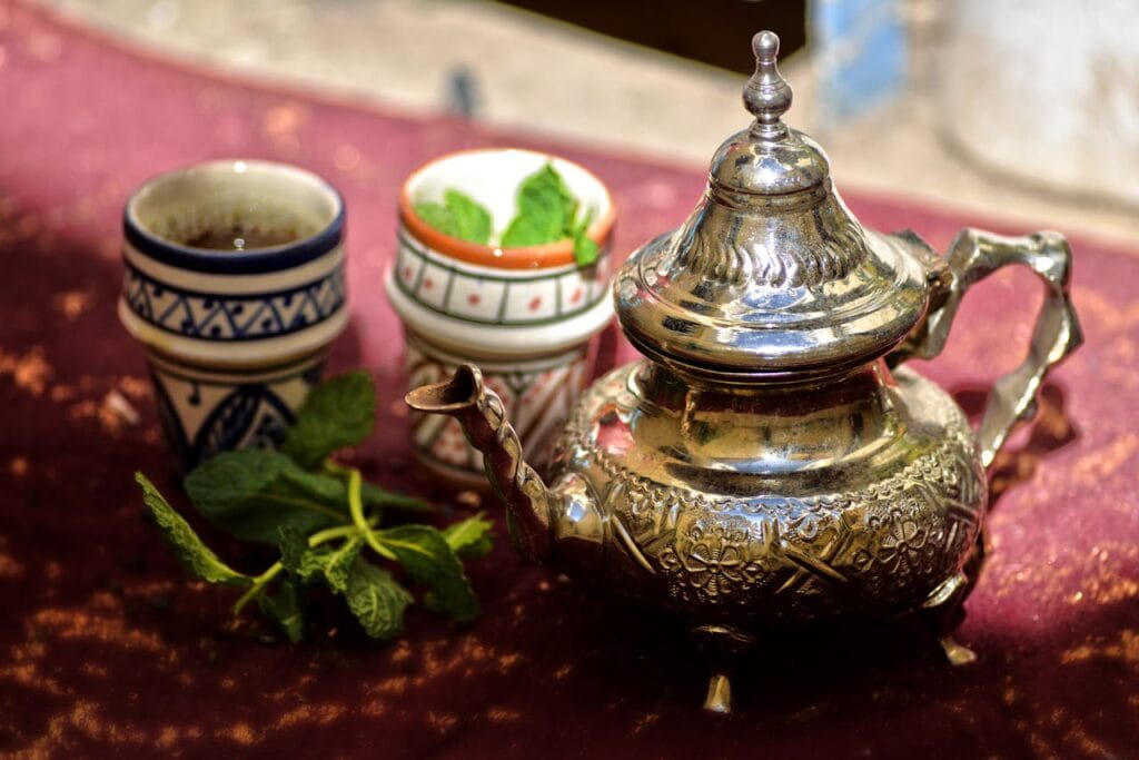A traditional Moroccan silver teapot with ceramic cups and fresh mint leaves on a table.