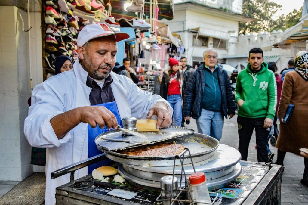 A Moroccan street food vendor preparing a traditional caliente in a busy medina with people walking in the background.