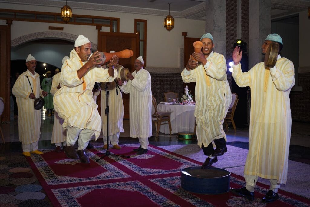 Moroccan traditional musicians performing a lively folk dance with drums and clay instruments during an indoor ceremony.