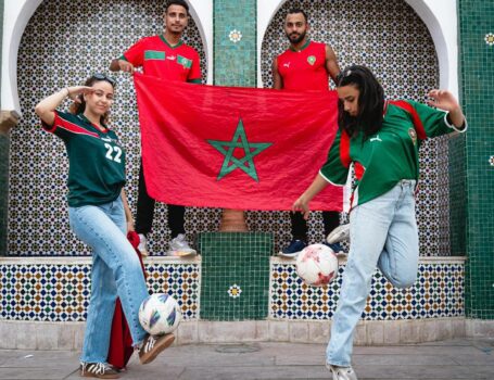 A group of Moroccan football supporters posing with the national flag while two young women juggle footballs in front of traditional zellij arches.