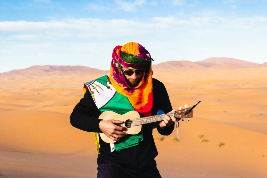 A tourist musician wearing a colorful desert scarf playing a small string instrument in the Sahara dunes near M’Hamid El Ghizlane.