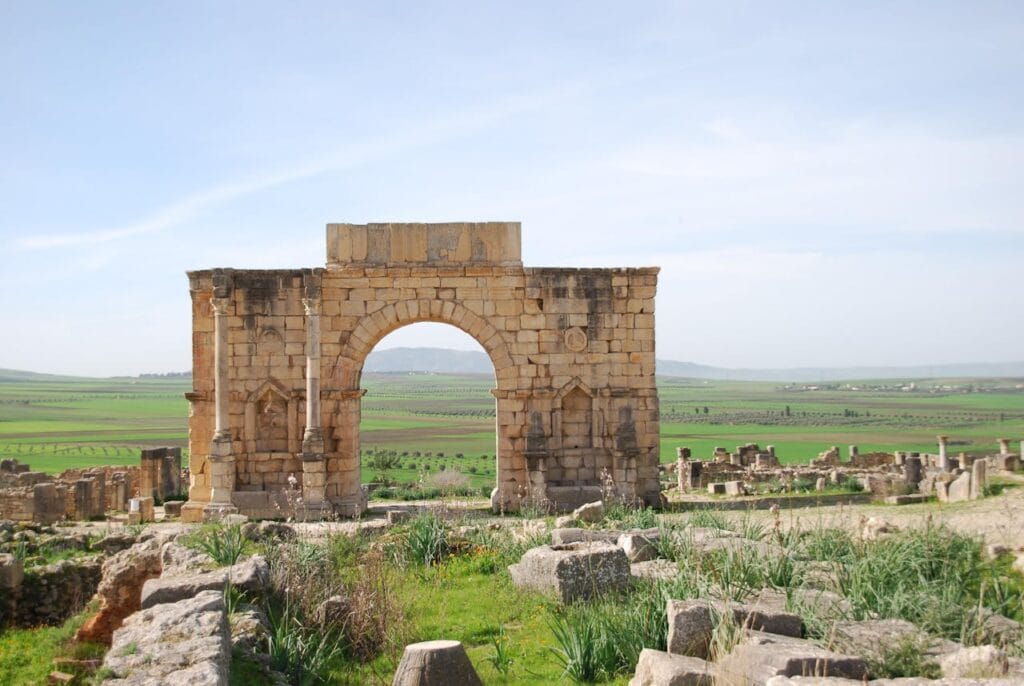 Roman arch at the ancient site of Volubilis in Morocco, surrounded by fields and archaeological ruins.