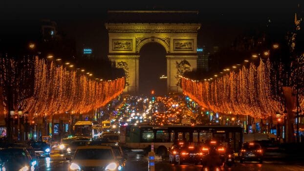 Night view of the Champs-Élysées in Paris with heavy traffic and illuminated decorations near the Arc de Triomphe.