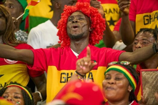 Ghana football supporters celebrating in the stands during an Africa Cup of Nations match, wearing national colors and waving flags.