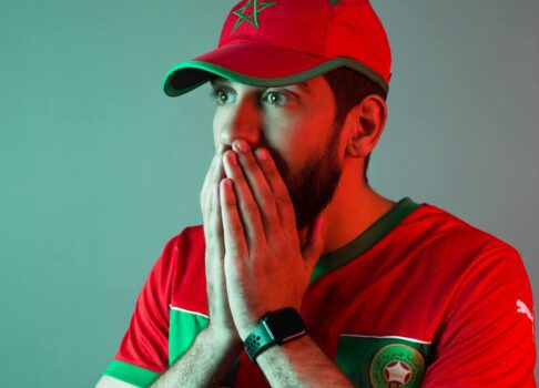 A Moroccan football fan wearing a red national team jersey and cap, holding his face in shock or anticipation.