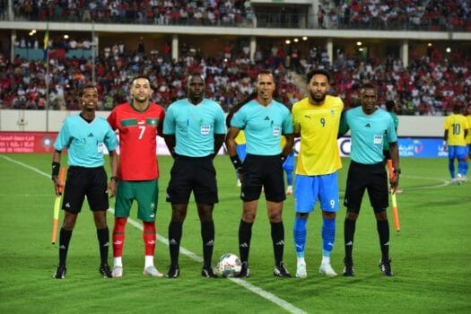 Pierre-Emerick Aubameyang and Hakim Ziyech posing with referees before a Morocco vs Gabon match, with a crowded stadium in the background.