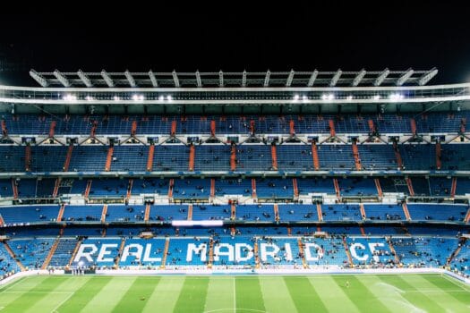 An illuminated Santiago Bernabéu Stadium with empty blue seating sections and the words “Real Madrid C.F.” displayed.